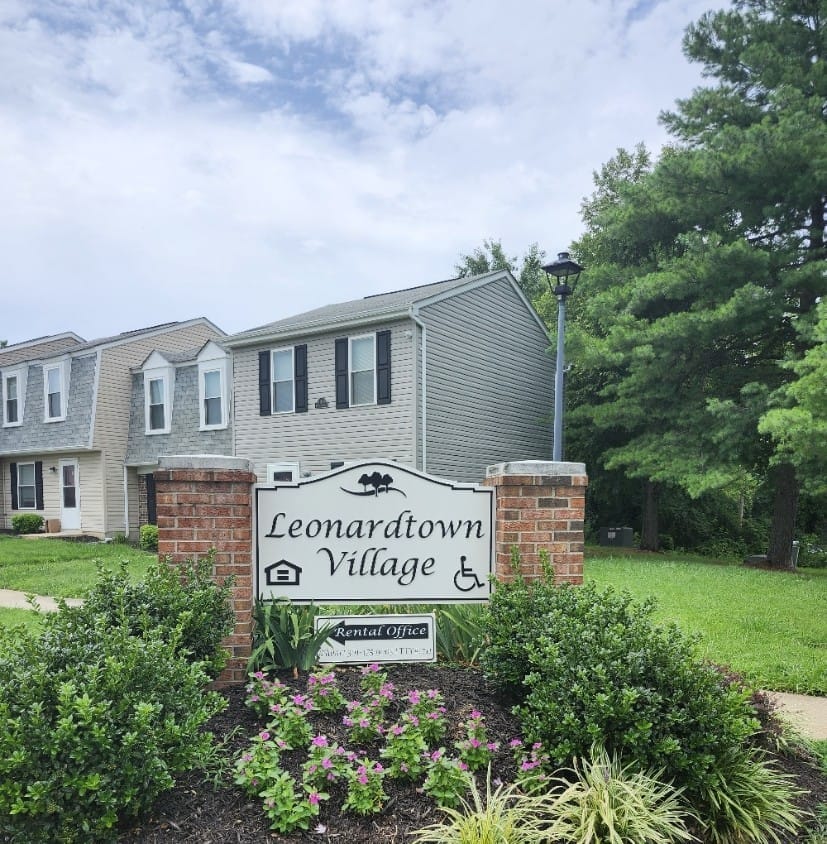 The Leonardtown Village monument sign on brick pillars, reading "Leonardtown Village" with a rental office plaque, a flower bed in front and a townhouse behind